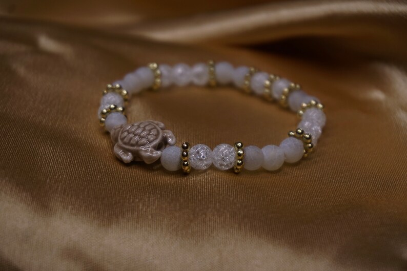 May include: A bracelet with white, crackled beads and gold-coloured accents, featuring a turtle charm. The turtle is light brown. The bracelet is displayed on gold-coloured fabric. The beads are approximately 0.8 cm in diameter.