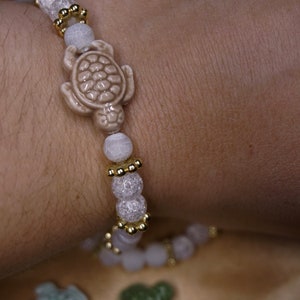 May include: A bracelet with a light beige turtle charm and light pink and white beads. Gold-coloured spacers add detail. The bracelet is designed to be worn on the wrist. The beads are measured in centimetres.