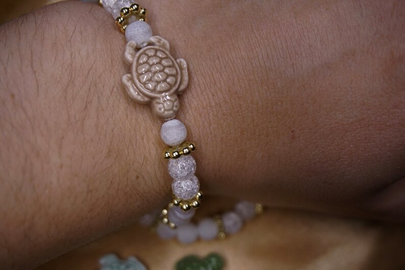 May include: A bracelet with a light beige turtle charm and light pink and white beads. Gold-coloured spacers add detail. The bracelet is designed to be worn on the wrist. The beads are measured in centimetres.