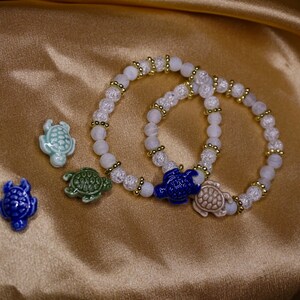 May include: Two beaded bracelets with white and gold details, each with a ceramic turtle charm. Three additional turtle charms in blue, green, and light blue are also shown. The jewellery is set against a gold satin background.