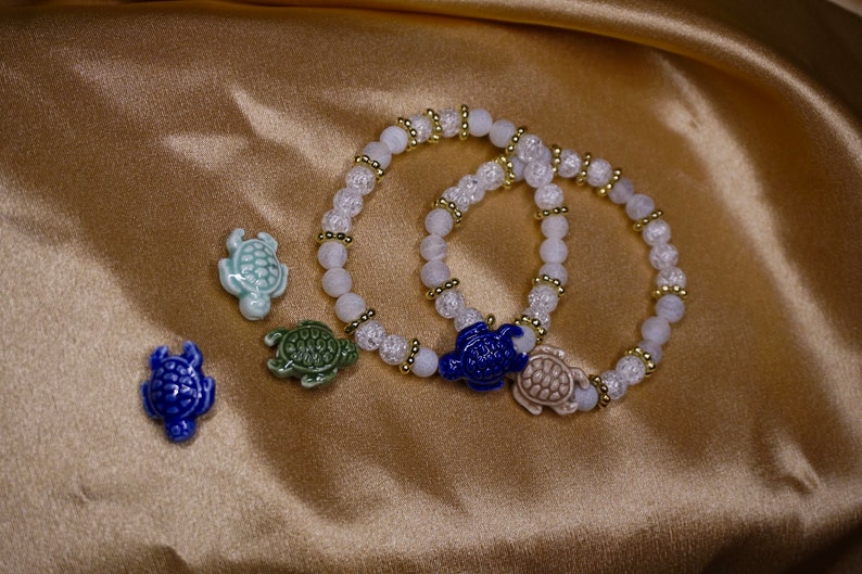 May include: Two beaded bracelets with white and gold details, each with a ceramic turtle charm. Three additional turtle charms in blue, green, and light blue are also shown. The jewellery is set against a gold satin background.