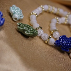 May include: A close-up of a bracelet and turtle charms. The bracelet features white beads with gold accents. The charms are ceramic turtles in blue, green, and off-white colours. The background is a golden fabric.