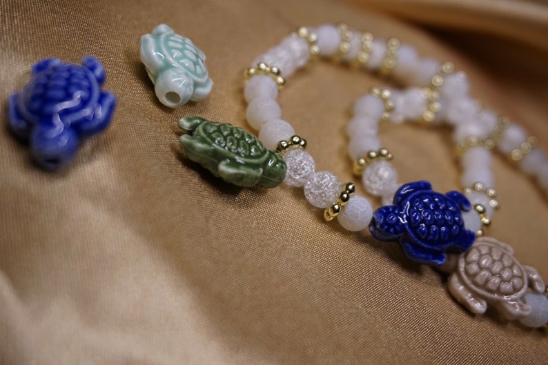 May include: A close-up of a bracelet and turtle charms. The bracelet features white beads with gold accents. The charms are ceramic turtles in blue, green, and off-white colours. The background is a golden fabric.