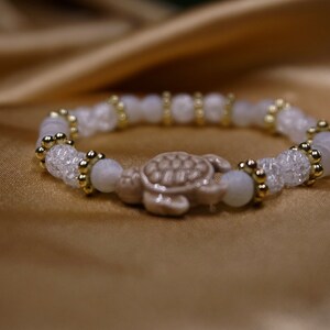May include: A white beaded bracelet with a turtle charm. The bracelet features clear, faceted beads, round white beads, and gold-coloured accents. The turtle charm is a light brown colour.