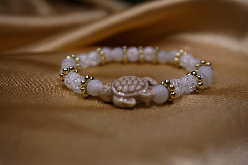 May include: A white beaded bracelet with a turtle charm. The bracelet features clear, faceted beads, round white beads, and gold-coloured accents. The turtle charm is a light brown colour.