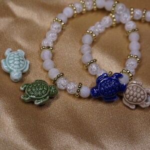 May include: Two bracelets with white and clear beads, and gold-coloured spacers. Each bracelet has a ceramic turtle charm. Additional turtle charms in blue, green, and light blue are scattered nearby.