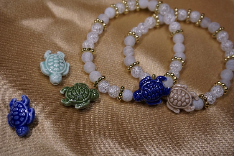 May include: Two bracelets with white and clear beads, and gold-coloured spacers. Each bracelet has a ceramic turtle charm. Additional turtle charms in blue, green, and light blue are scattered nearby.