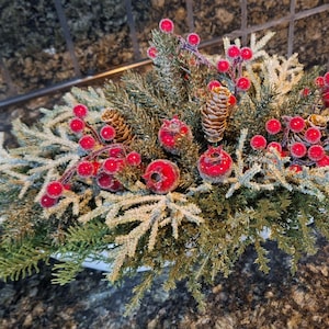 May include: A festive Christmas centerpiece featuring evergreen branches, frosted white sprigs, red berries, and pinecones. The arrangement is set in a white container, perfect for holiday decor.