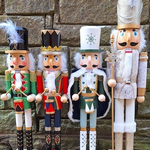 Puede incluir: Cuatro cascanueces de madera decorativos en fila. Cada figura tiene un diseño único con uniformes y sombreros coloridos, detalles como plumas y adornos dorados. Los cascanueces están pintados en rojo, verde y dorado, y están sobre una superficie de piedra.