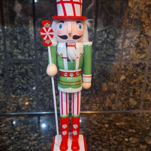 May include: A decorative wooden nutcracker figurine. The figurine is dressed in a green jacket, red boots, and a tall white hat with red stripes. It holds a candy cane-shaped staff and stands on a red and white base. A festive holiday decoration.