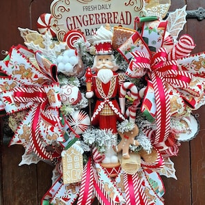 May include: A festive Christmas wreath with a gingerbread theme. It features a red and white color scheme with ribbons, candy canes, gingerbread men, and a nutcracker. The sign reads "Fresh Daily Gingerbread Bar".