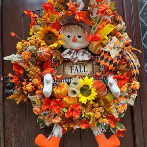 May include: A seasonal wreath with a scarecrow motif. It features a scarecrow head with a brown hat, sunflowers, pumpkins, and a sign reading "Welcome Fall". The colour scheme is predominantly orange, yellow, and brown, perfect for autumn.