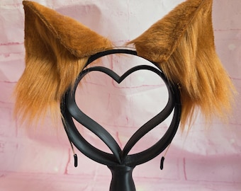 May include: A pair of brown faux fur cat ears attached to a black headband. The ears are upright and fluffy, with a darker brown interior. The headband is supported by a black heart-shaped stand, creating a whimsical accessory.