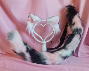 May include: A pink and black cat tail with a white tip, attached to a black strap. The tail is displayed on a pink surface with a white heart-shaped stand and a pair of pink and white cat ears.