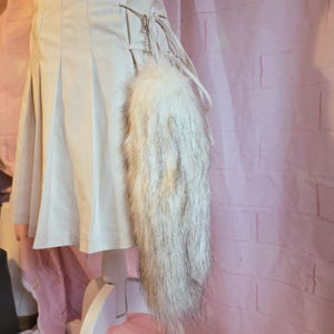 K&ouml;nnte beinhalten: Ein wei&szlig;-grauer Kunstfellschwanz mit einer silbernen Kette und Lederb&auml;ndern. Der Schwanz ist an einem cremefarbenen Rock befestigt. Der Hintergrund ist eine rosa Backsteinmauer.