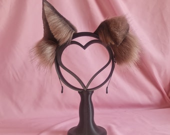 May include: A brown and tan faux fur headband with attached ears, displayed on a black stand with a heart-shaped design. The ears have a fluffy texture and are designed to resemble animal ears. The background is a solid pink colour.