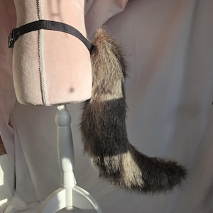 May include: A faux fur tail in shades of brown and gray, attached to a black belt with a buckle. The tail is displayed on a pink mannequin, showcasing its fluffy texture and shape.