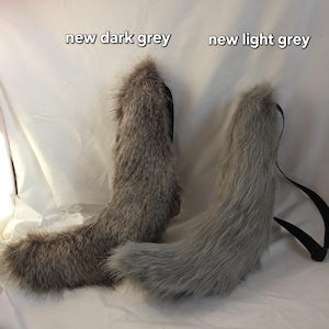 May include: Two faux fur tails, one dark grey and one light grey, with black straps. The tails are fluffy and have a curved shape. Text at the top reads "new dark grey" and "new light grey".