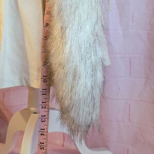 K&ouml;nnte beinhalten: Ein wei&szlig;-grauer Kunstfellbesatz, ca. 25 cm lang, wird mit einem rosa Ma&szlig;band gemessen. Der Besatz ist an einem wei&szlig;en Kleidungsst&uuml;ck befestigt. Der Hintergrund ist eine rosa Backsteinmauer.