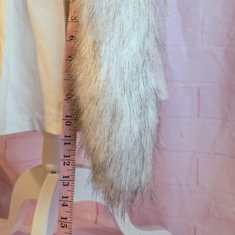 K&ouml;nnte beinhalten: Ein wei&szlig;-grauer Kunstfellbesatz, ca. 25 cm lang, wird mit einem rosa Ma&szlig;band gemessen. Der Besatz ist an einem wei&szlig;en Kleidungsst&uuml;ck befestigt. Der Hintergrund ist eine rosa Backsteinmauer.