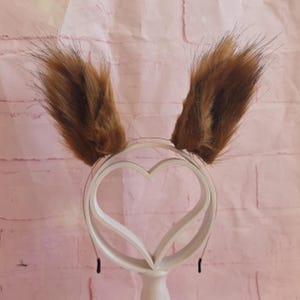 May include: A headband with fluffy, brown faux fur ears. The ears are attached to a white, heart-shaped frame. The headband is designed to be worn on the head and is suitable for cosplay or costume parties.