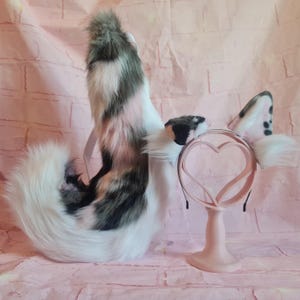 Puede incluir: Una cola de gato rosa, blanca y gris con un par de orejas de gato a juego en una diadema rosa. Las orejas tienen manchas negras y se muestran en un soporte rosa.