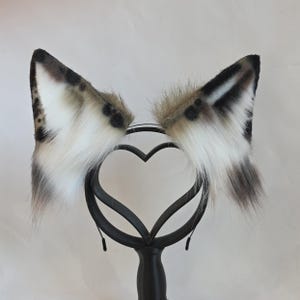 May include: Faux fur cat ears in shades of white, brown, and black, attached to a black headband. The ears have a spotted pattern and are fluffy. The headband is displayed on a black stand with a heart-shaped design.
