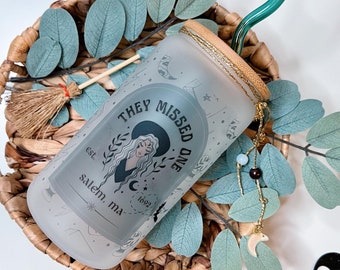 May include: A frosted glass tumbler with a black and white design that says "They Missed One est. 1692 Salem, MA". The tumbler is in a wicker basket with green leaves and a green glass straw.