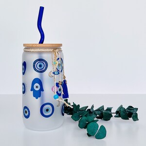 May include: A frosted glass water bottle with a blue and white evil eye design and a blue glass straw. The bottle has a wooden lid and a gold tassel charm.