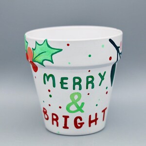 May include: White ceramic flower pot with green holly leaves and red berries painted around the top. The words "Merry & Bright" are painted in green and red on the front.