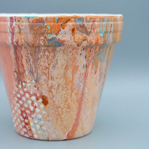 Pode incluir: Um vaso de terracota pintado com um design marmorizado em tons de laranja, pêssego e branco. O vaso tem um padrão texturizado de pontos brancos em um lado.