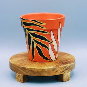 May include: A terracotta flower pot with a hand-painted design of black and white leaves on an orange background. The pot is sitting on a wooden stand with three legs.