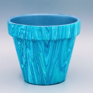 May include: A small, blue and turquoise marbled ceramic flower pot. The pot has a flared shape with a slightly wider rim and a smooth, glossy finish. The interior is a solid, muted blue.