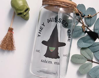 May include: A clear glass reusable water bottle with a bamboo lid and a straw. The bottle has a black and white graphic of a witch with the text "They Missed One est. 1692 Salem, Ma".