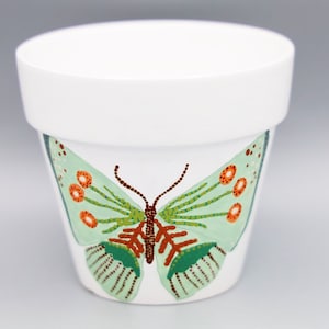 May include: A white ceramic flower pot with a green butterfly painted on the front. The butterfly has orange and brown accents.