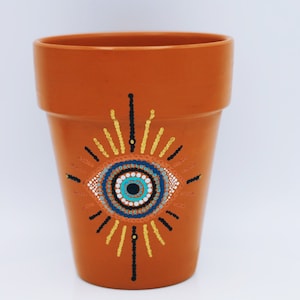 May include: Terracotta flower pot with a hand-painted evil eye design. The eye is blue with white and black accents, surrounded by a yellow sunburst.