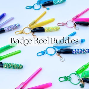 May include: A collection of colourful, jeweled pens with keychains. The pens are decorated with rhinestones and have a variety of colours, including pink, blue, green, and yellow. The keychains are attached to the pens and have a variety of designs, including a smiley face and a star. The text "Badge Reel Buddies" is written in the centre of the image.
