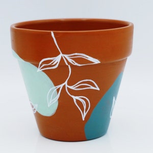 Puede incluir: Maceta de terracota con un diseño pintado a mano que presenta formas abstractas blancas y hojas sobre un fondo marrón, azul verdoso y verde claro.