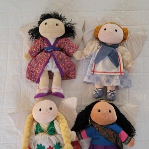 May include: Four handmade cloth dolls with smiling faces and various outfits. One doll wears a red floral robe, another a blue and white dress, a third a green and red dress, and the last a blue top and purple pants. Each doll has attached wings.