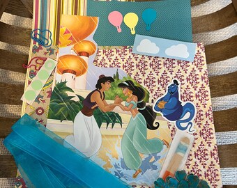 Peut inclure: Un kit de scrapbooking comprenant une illustration centrale d'Aladdin et Jasmine, une découpe du Génie bleu et des éléments décoratifs dont des bandes de papier, des montgolfières en papier, des nuages, un ruban bleu sarcelle et des fleurs.