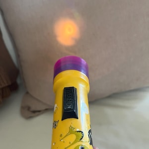 May include: A yellow Eveready flashlight with a purple lens cap and a black sliding power switch. The body features black and green graphic illustrations of cats and the text W. The flashlight emits a warm orange light beam from the front lens.