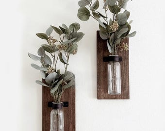 May include: Two wall-mounted decor pieces, each consisting of a rectangular dark brown wooden board with a clear glass bottle attached by a black metal bracket, holding stems of green eucalyptus leaves with small seed clusters.