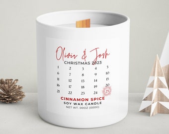 May include: A white cylindrical soy wax candle with a wooden wick. The label features the text "Olivia & Josh", "CHRISTMAS 2023", a calendar grid with the 25th highlighted, and the text "CINNAMON SPICE SOY WAX CANDLE" with "NET WT. 00OZ (000G)".