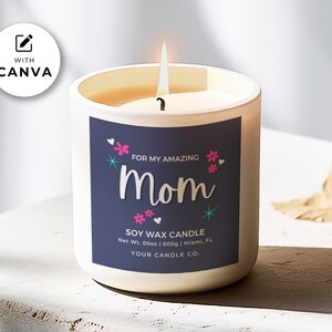 May include: A white soy wax candle with a blue label that reads "For My Amazing Mom" with pink flowers and stars. The candle is on a white surface with a brown leaf in the background.