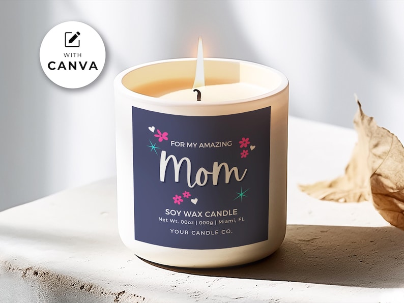 May include: A white soy wax candle with a blue label that reads "For My Amazing Mom" with pink flowers and stars. The candle is on a white surface with a brown leaf in the background.