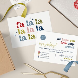 May include: A flat lay featuring holiday-themed stationery. Two cards display the text "fa la la" and "We hope you love your order!" with a festive design. A brown box, ribbon, and scissors are also present.