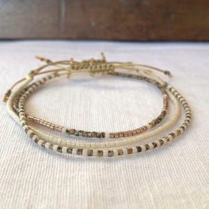 May include: Three delicate beaded bracelets in shades of white, brown, and gold. The bracelets are layered on a white fabric background.