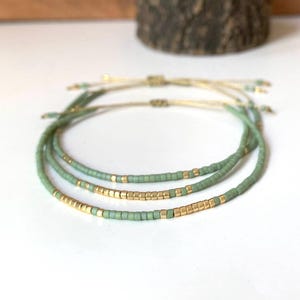 May include: Three delicate beaded bracelets. Each bracelet features a combination of small, light green beads and gold-coloured square beads. The bracelets are strung on a thin, beige cord, with adjustable closures. The bracelets are displayed on a white surface.