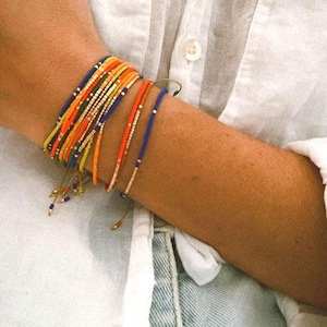 May include: A collection of colorful beaded bracelets in shades of blue, orange, yellow, and gold. The bracelets are stacked on a wrist, creating a vibrant and stylish accessory. The bracelets are set against a white shirt and denim.