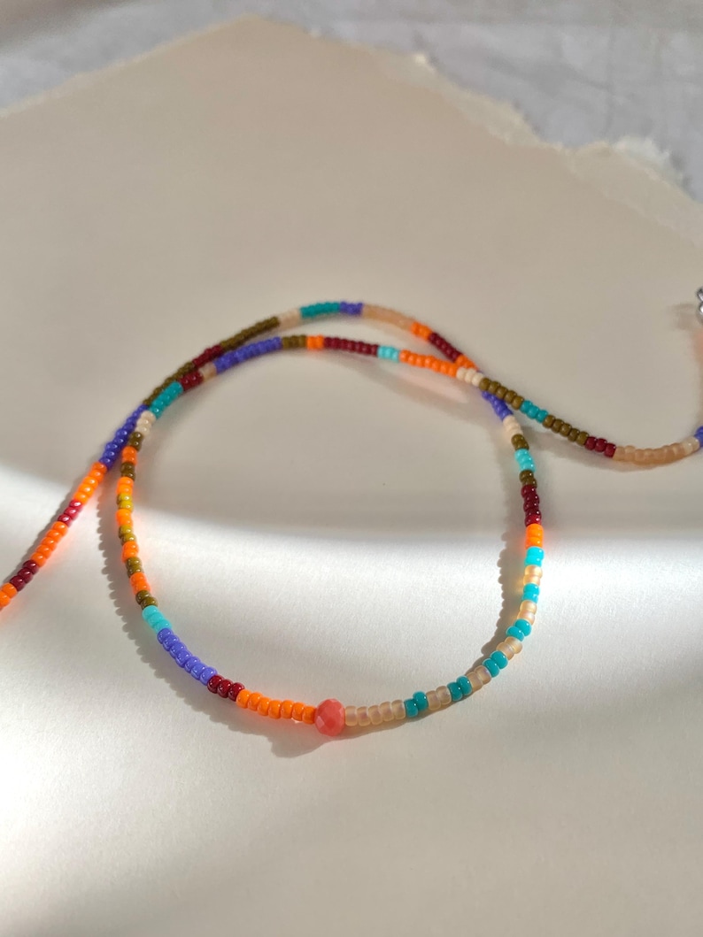 A multi-colored beaded necklace with a single larger coral colored bead in the center. The necklace is made of small, round beads in shades of brown, orange, red, purple, blue, and turquoise.